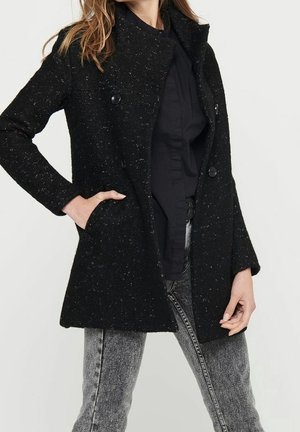 Femme portant un manteau noir à pois à double boutonnage sur un chemisier noir et un jean gris délavé à l'acide, avec une main dans la poche.