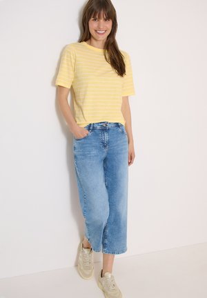 Kvinde smiler, iført gulstribet t-shirt, lyseblå vid-benet cropped jeans, beige sneakers, står mod en hvid væg.