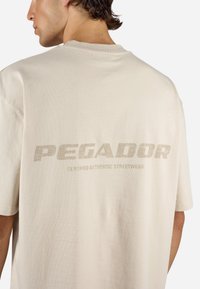 Beige oversized t-skjorte med en teksturert trykk med ordet "PEGADOR" og "CERTIFIED AUTHENTIC STREETWEAR" i lys brun på baksiden.