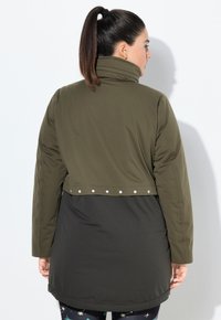Mujer con coleta oscura que lleva una chaqueta acolchada bi-tono en oliva y negro con puntos reflectantes en el dobladillo trasero, de pie contra una pared blanca.