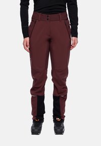 Person, der eine dunkelrote Outdoor-Hose mit schwarzen Knie- und Wadenpatches, schwarze Stiefel und ein langärmliges schwarzes Oberteil trägt, gerade steht.