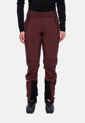 Persona che indossa pantaloni outdoor rosso scuro con toppe nere sulle ginocchia e sui polpacci, stivali neri e una maglietta a maniche lunghe nera, in piedi dritta.