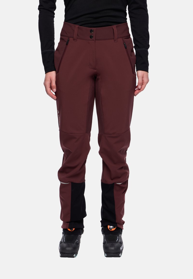 Person, der eine dunkelrote Outdoor-Hose mit schwarzen Knie- und Wadenpatches, schwarze Stiefel und ein langärmliges schwarzes Oberteil trägt, gerade steht.