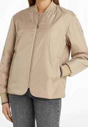 Femme portant une veste bomber beige zippée avec des poignets côtelés et un jean en denim gris, main dans la poche sur fond blanc.