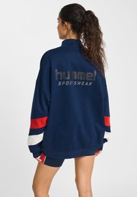 Felpa oversize blu navy con maniche a strisce rosse, bianche e blu, caratterizzata da un audace logo "hummel SPORTSWEAR" sulla schiena.
