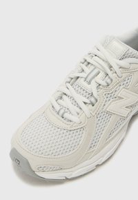 Chaussure de sport légère blanche avec une tige en mesh, un col rembourré et des accents synthétiques texturés ; dotée de lacets blancs et d'une semelle grise.
