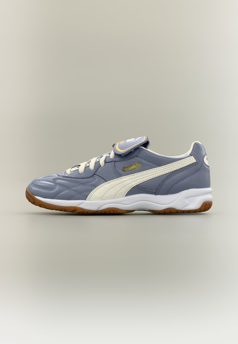 Γκρι και λευκό αθλητικό παπούτσι Puma με σόλα από καουτσούκ, μπροστινή δέση με κορδόνια και επωνυμία στο καπάκι της γλώσσας, εμφανιζόμενο σε πλευρικό προφίλ σε ουδέτερο φόντο.