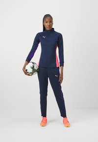 Atlétický outfit Puma v námorníckej modrej farbe s ružovými a fialovými akcentmi, obsahujúci zipsovú dlhú mikinu a zodpovedajúce nohavice. Hráč drží futbalovú loptu.