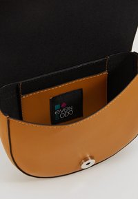 Jasnobrązowa torebka z ekoskóry o zaokrąglonym kształcie, czarnym wnętrzu i srebrnym zatrzaskiem; wewnątrz znajduje się metka marki.