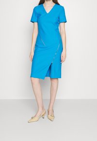 Modré šaty s krátkými rukávy, výstřihem do V a diagonálním zipem, s délkou ke kolenům a předním rozparkem. Outfit doplňují béžové lodičky.