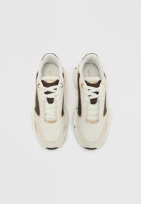 Tennarit, joissa on valkoinen nahkainen päällinen, ruskeat mokkanahkaiset yksityiskohdat, kullanväriset metalliosat ja paksu musta kumipohja pyöreällä muotoilulla.