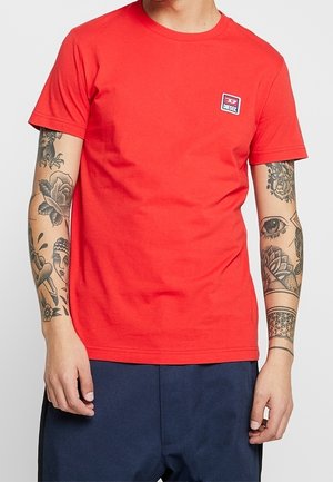 Mężczyzna w czerwonym t-shircie Diesel z małym logo na piersi i widocznymi tatuażami na obu przedramionach, w ciemnoniebieskich spodniach.