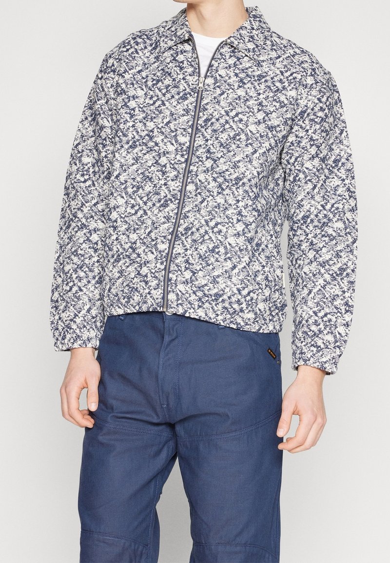 Quiltet jakke med et blåt og hvidt teksturmønster, lynlåslukning og krave, kombineret med marineblå bukser.