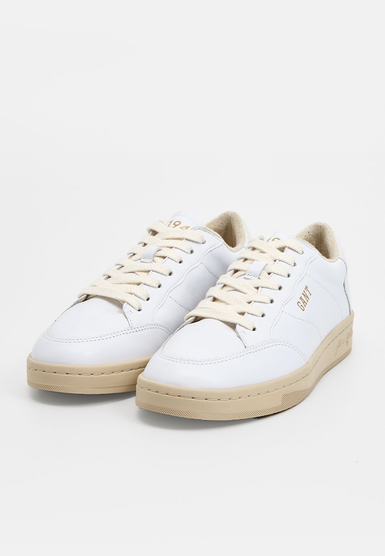 Zapatillas de piel blancas con suela y cordones en beige. Presentan un logotipo dorado y un diseño clásico de caña baja. Superficie texturada y detalle de costura.