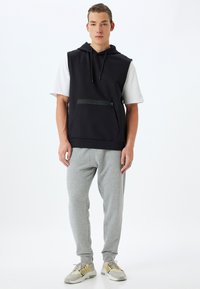Sudadera sin mangas negra con un bolsillo frontal, combinada con pantalones de chándal gris claro. La sudadera cuenta con un cordón y un acento con cremallera.