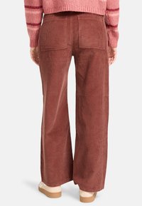 Pantaloni a gamba larga in corduroy marrone con due tasche posteriori e una superficie texturizzata, mostrati dal retro con un maglione rosa texturizzato.