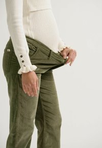 Olivgrüne Cordhosen mit lockerer Passform und Reißverschluss, getragen zu einem eng anliegenden weißen Rippsweater mit Rüschensäumen und Knopfakzenten.