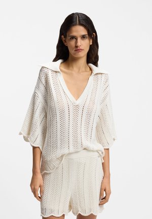 Vrouw met een witte gebreide V-hals top met kraag en bijpassende shorts met hoge taille, staand tegen een effen achtergrond.