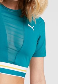 Teal atletisk crop top med korta ärmar, nätpanel och randig band i vitt, gult och teal vid fållen. Puma-logotyp på bröstet.