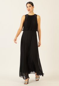 Schwarzes, plissiertes Maxikleid mit ärmellosem Design, transparentem Overlay am Oberteil und fließendem Rock. Kombiniert mit verzierten offenen Schuhen.