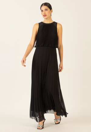 Schwarzes, plissiertes Maxikleid mit ärmellosem Design, transparentem Overlay am Oberteil und fließendem Rock. Kombiniert mit verzierten offenen Schuhen.