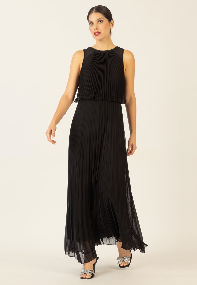 Schwarzes, plissiertes Maxikleid mit ärmellosem Design, transparentem Overlay am Oberteil und fließendem Rock. Kombiniert mit verzierten offenen Schuhen.