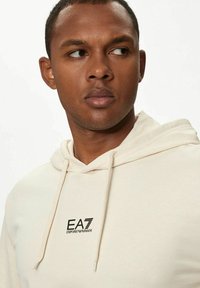 Felpa beige con cappuccio e coulisse, caratterizzata da un logo EA7 Emporio Armani nero sul petto. Tessuto morbido, vestibilità rilassata e tasca a marsupio.