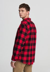 Giovane uomo di profilo che indossa una camicia a quadri rossi e neri e pantaloni beige su uno sfondo chiaro e uniforme.