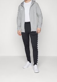 Sweat à capuche gris zippé sur un T-shirt blanc, associé à un pantalon foncé avec des bandes latérales. Des baskets blanches complètent la tenue.