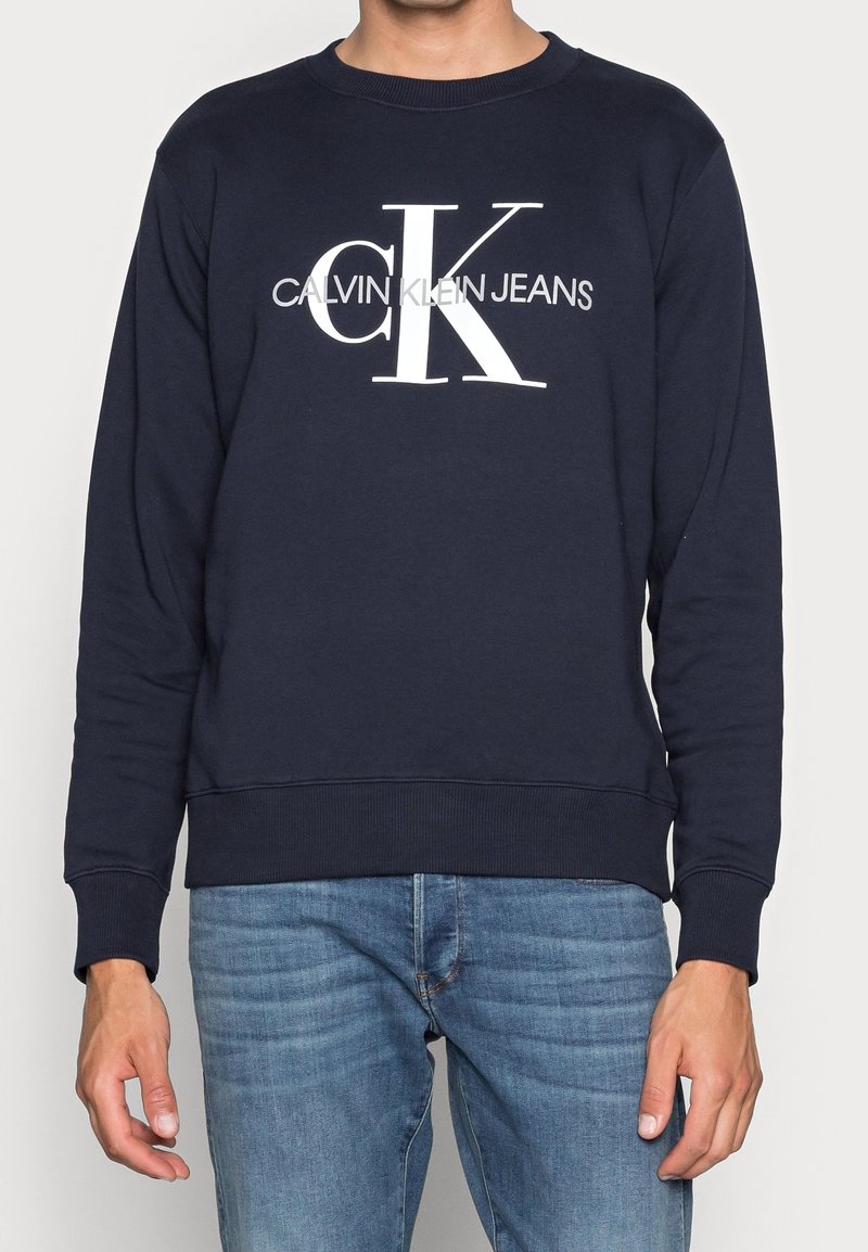 Marinblå bomullströja med ribbad rund hals, långa ärmar och vit tryckt logotyp "CALVIN KLEIN JEANS" tydligt synlig.