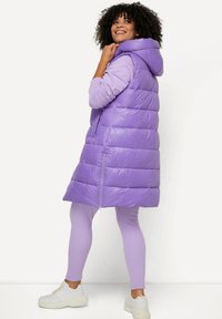 Gilet matelassé violet avec capuche, présentant une texture brillante, un motif matelassé horizontal et des poches latérales. Porté sur un haut violet assorti et des leggings.