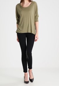 Olivově zelený volný top s křídly, kombinovaný s černými slim-fit legínami a černými vysokými podpatky. Hladká textilie.