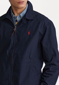 Navyblaues Windbreaker-Jacke mit einem Reißverschluss vorne, einem Punktkragen und roten Logo-Akzenten. Ausgestattet mit zwei Vordertaschen und einem leichten Material.