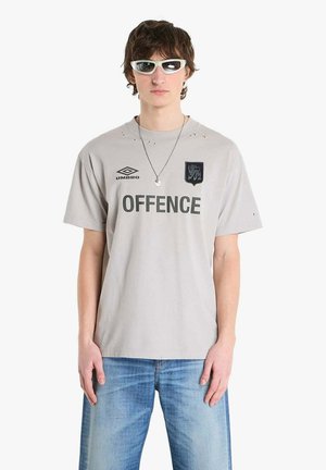 Giovane uomo che indossa occhiali da sole bianchi, una maglietta grigio chiaro con la scritta "OFFENCE" e jeans blu, in piedi davanti a uno sfondo bianco semplice.