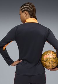 Maglia sportiva nera a maniche lunghe con accenti arancioni sul collo e sulle maniche, abbinata a un pallone da calcio dorato con un motivo e un logo neri.