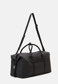 Le sac de sport en toile noire présente un motif texturé, deux poignées supérieures, une bandoulière réglable et un accent de logo sur le côté.