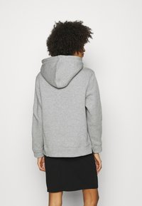 Person med lockigt hår iförd en grå hoodie och svart kjol, vänd bort från en enkel vit bakgrund.