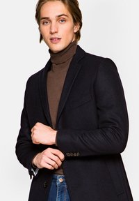 Granatowa wełniana marynarka z guzikiem, z klapami i kieszenią na piersi, nałożona na brązowy sweter z golfem.