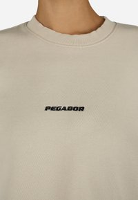 Bežs sporta krekls ar ribotu apkakli un melnu izšūtu "PEGADOR" logo uz krūtīm. Gluda auduma tekstūra.