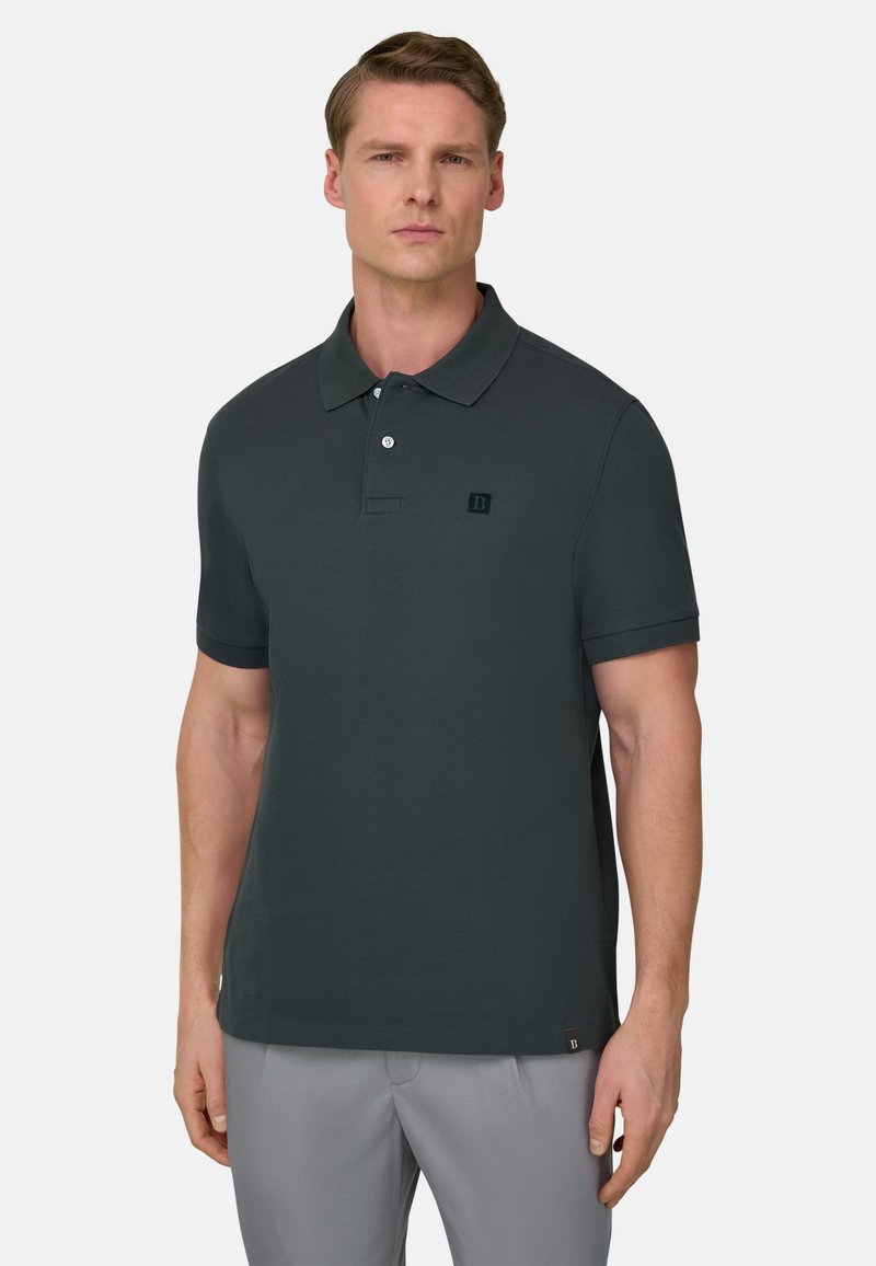 Homme portant un polo à manches courtes vert foncé avec des boutons, debout devant un fond uni clair.