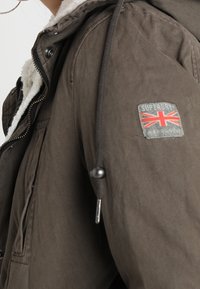 Chaqueta marrón texturizada con capucha y forro de pelaje sintético. Presenta un parche de logo con la bandera del Reino Unido en la manga y cordones ajustables.