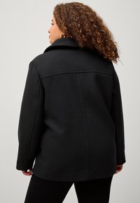 Cappotto nero in misto lana, spesso, con collo a scialle, vestibilità oversize e cuciture in vista. Tessuto liscio, lunghezza a metà fianchi e maniche lunghe.