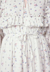 Valkoinen kukkamekko, jossa on röyhelökoristeita ja pieniä sinisiä ja vaaleanpunaisia kukkia. Valmistettu teksturoidusta kankaasta, jossa on sidottava vyö.