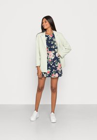 Blazer verde-claro sobre um vestido floral azul-marinho com saia rodada. Ténis brancos completam o look. Tecidos suaves com padrões variados.