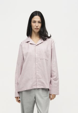 Femme aux longs cheveux foncés portant une chemise ample rose et blanche rayée à boutons et un pantalon gris clair, debout devant un fond blanc uni.