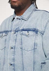 Valkoinen sininen farkkutakki, jossa on kaksi rintataskua, nappisulkijat ja kontrastivärinen ompelu. Kangas on pehmeä ja teksturoitu.