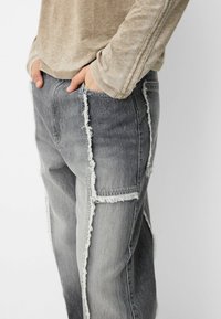 Šedé džínové kalhoty z denimu s opotřebovanými, třepenými švy, volným střihem a bočními kapsami. Kombinovány s lehkým béžovým topem s dlouhým rukávem.