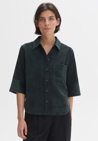 Dark green corduroy shirt with a collar, short sleeves, and a front pocket, featuring a soft texture and button closure.