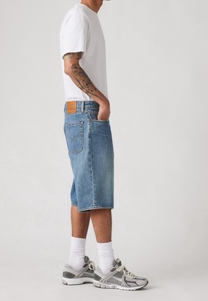 Man draagt wit T-shirt, knielange blauwe denim shorts, witte sokken en grijze sneakers, staat met hand in zak.