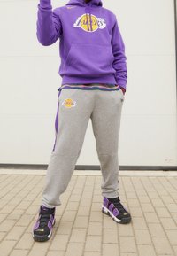 Fioletowa bluza z kapturem z logo Los Angeles Lakers, szare spodnie dresowe z logo oraz czarne sneakersy z fioletowymi akcentami na powierzchni z cegieł.